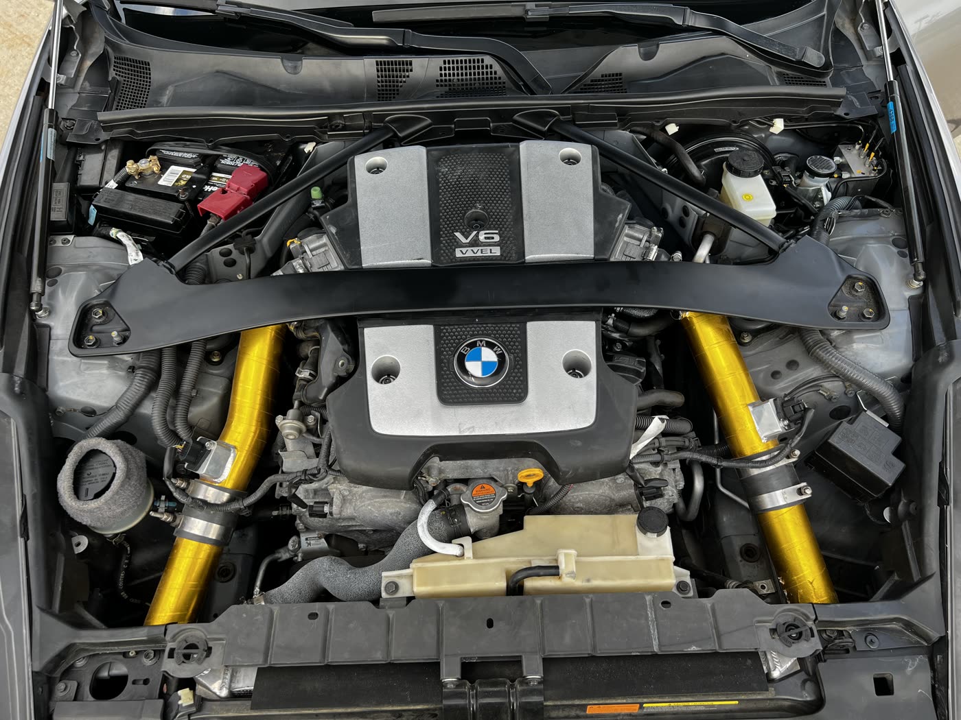 The 370Z's V6 engine bay with a strut brace, gold heat-wrapped intake pipes and a BMW roundel stuck on the engine cover