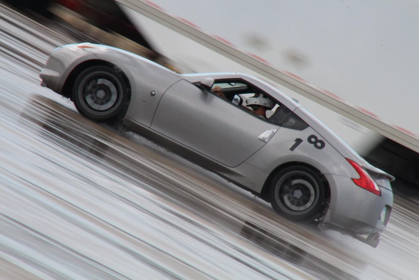 A silver 370Z, number 18, sliding through a wet autocross course, the background blurred by the camera pan