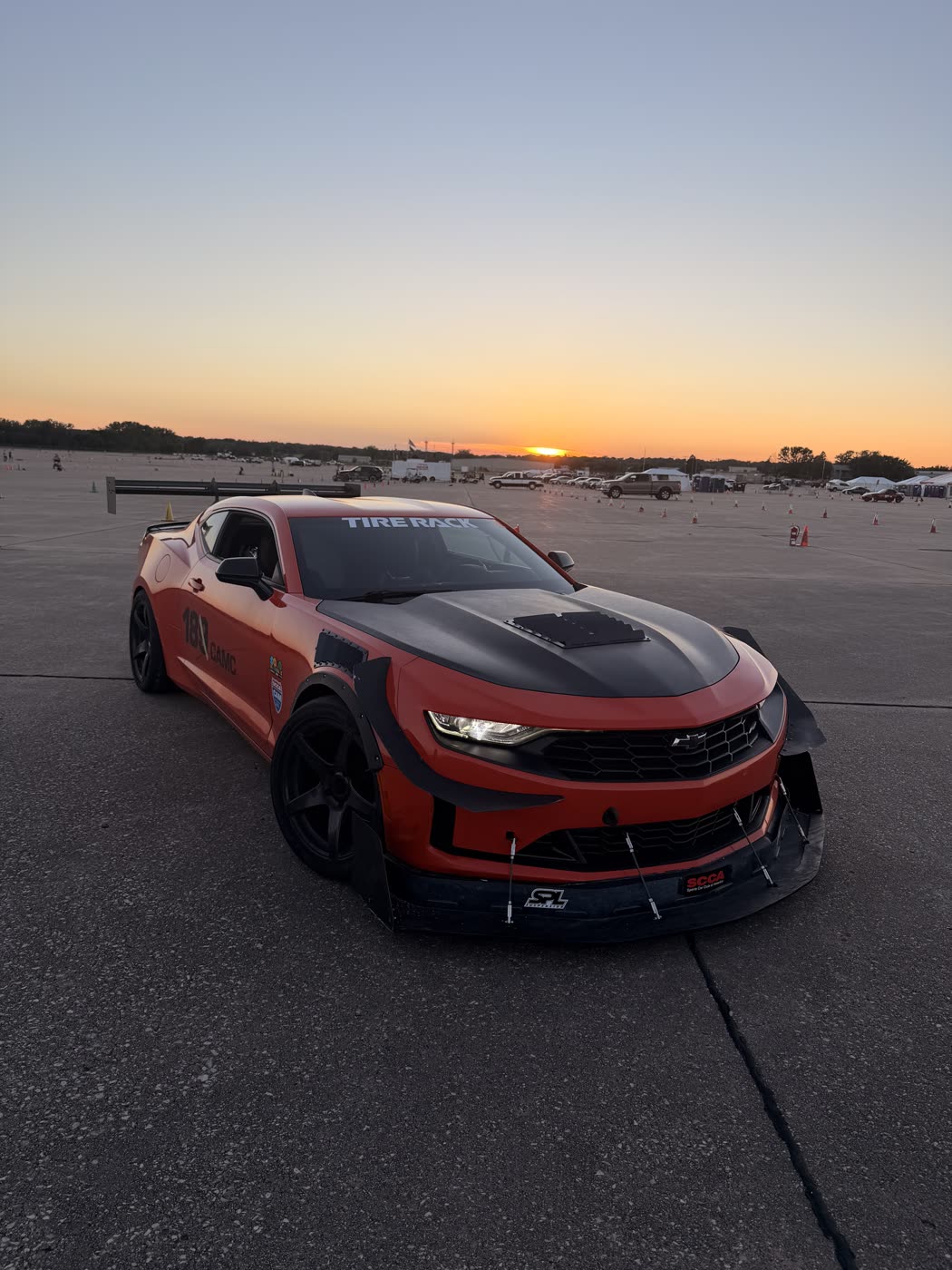 An orange Camaro with a wide front splitter, fender flares and a rear wing on an autocross lot at sunset