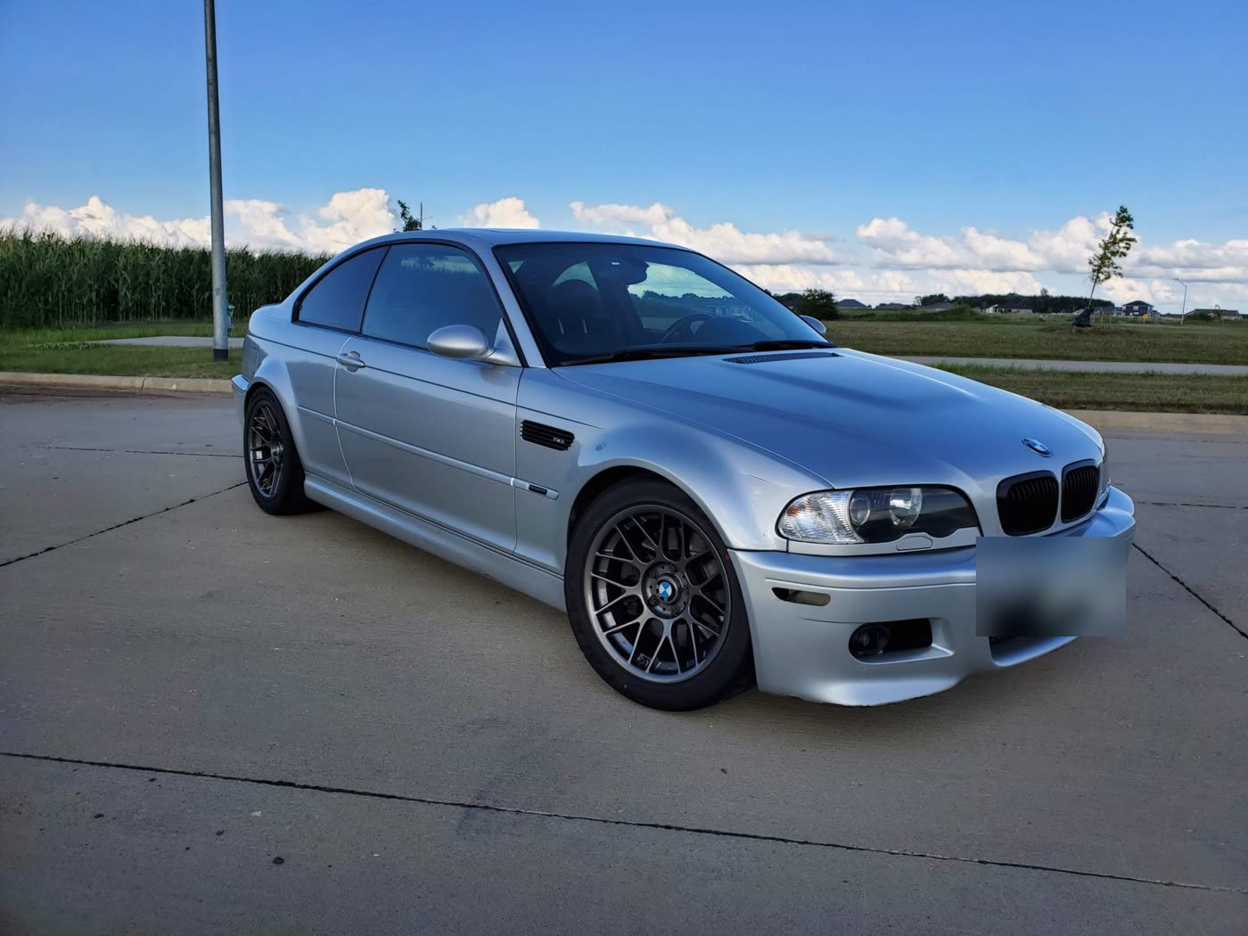 A silver E46 BMW M3 coupe on gray aftermarket wheels, parked beside a cornfield