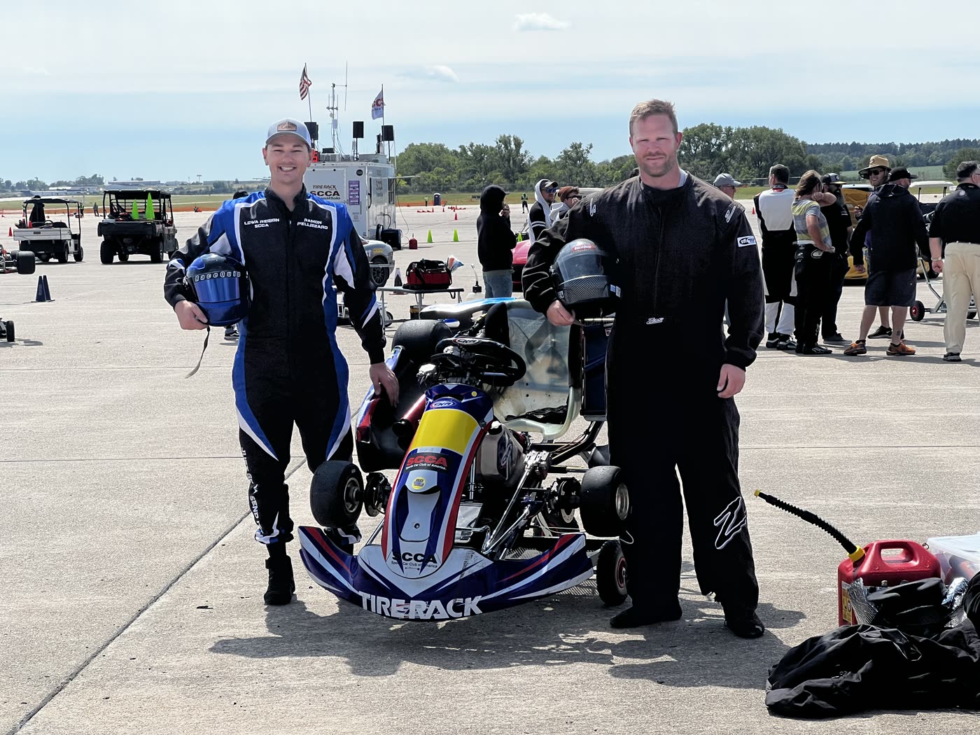 Ramon and Dan in racing suits, holding helmets, standing either side of a kart at an SCCA event