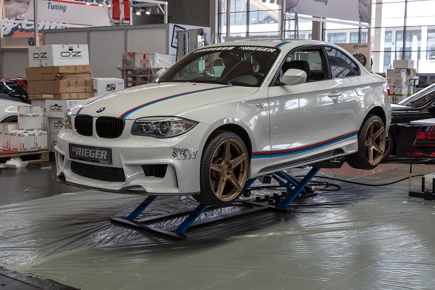 A modified BMW 1 Series E82 coupe with a body kit and bronze wheels, raised on a display lift at a tuning show
