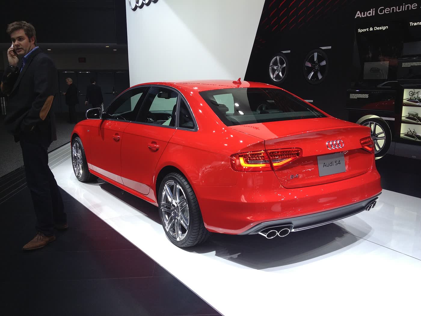 The rear three-quarter view of a red Audi S4 sedan on a show-floor stand, showing its four doors, the trunk lid with an S4 plate, and the quad exhaust tips under the bumper