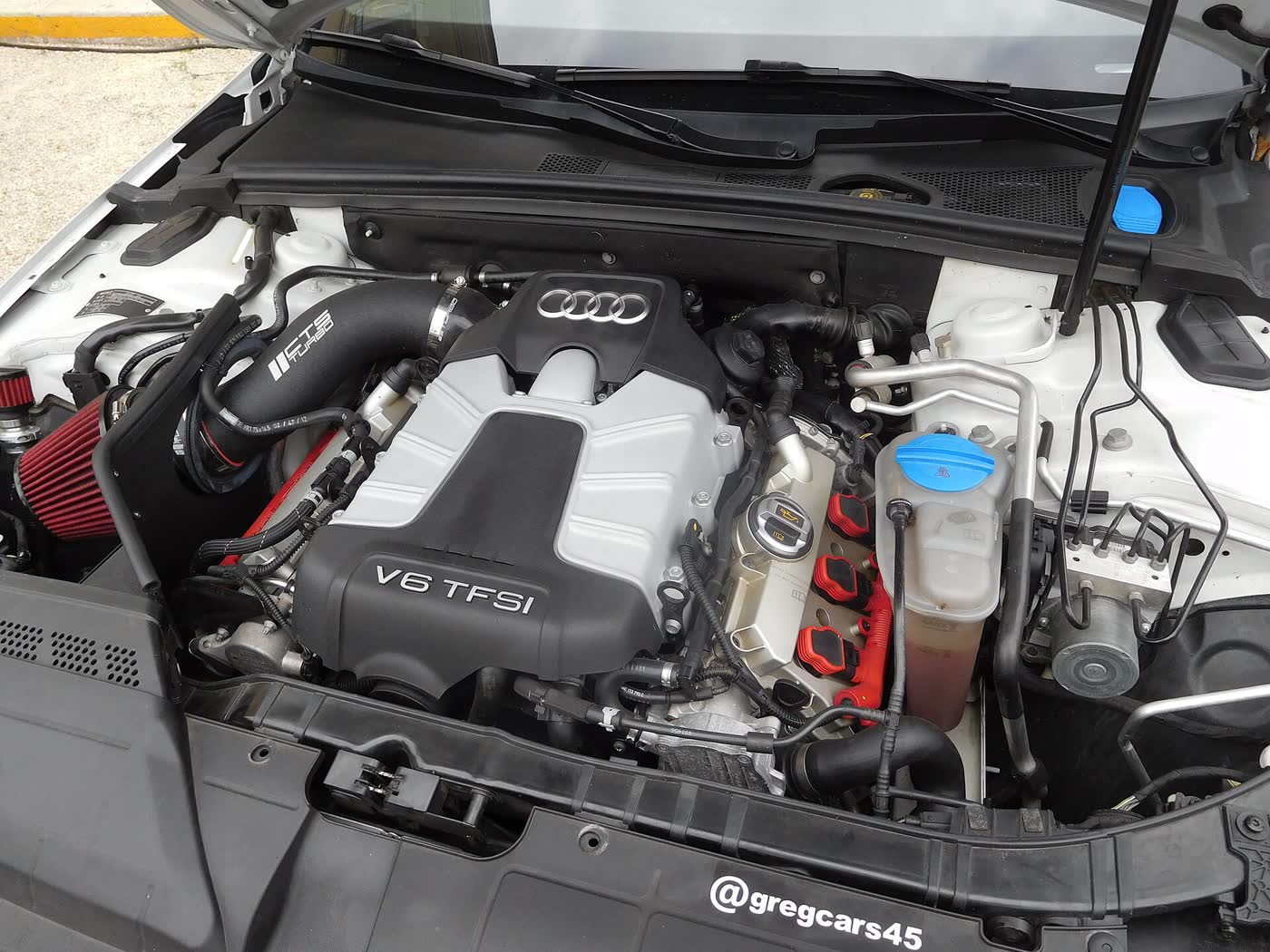 The engine bay of a white Audi S4 sedan with the hood raised, showing the V6 TFSI engine cover, red ignition coils and an aftermarket air intake with a red filter