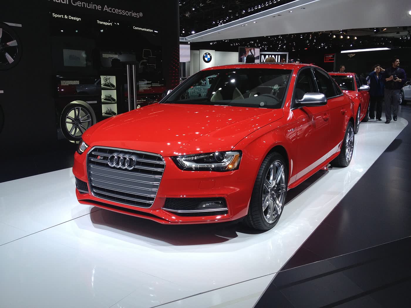 A red Audi S4 sedan on a white show-floor stand, front three-quarter view, with its single-frame grille, LED-lit headlights and polished multi-spoke wheels, other cars and visitors behind it