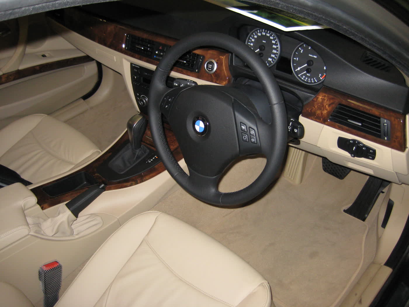 The front of a BMW E90 cabin seen through the open driver's door: cream leather seats, a wood-trimmed dashboard, a three-spoke steering wheel on the right-hand side and no screen on top of the dash