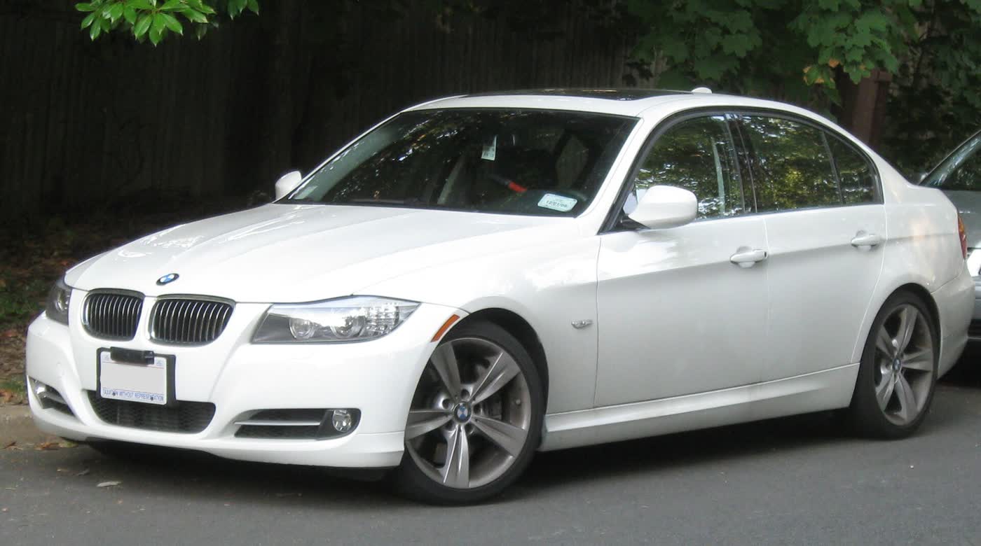 A white BMW 335i sedan with the later headlight and bumper styling, front three-quarter view, parked at a curb with trees behind it