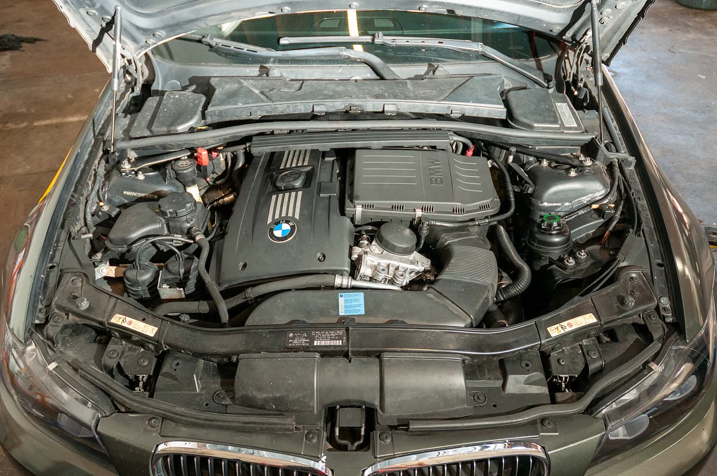 The open engine bay of a grey BMW 3 Series: a ribbed black engine cover with a BMW roundel, a black airbox to its right, and a silver oil filter housing with a round cap in front of the airbox