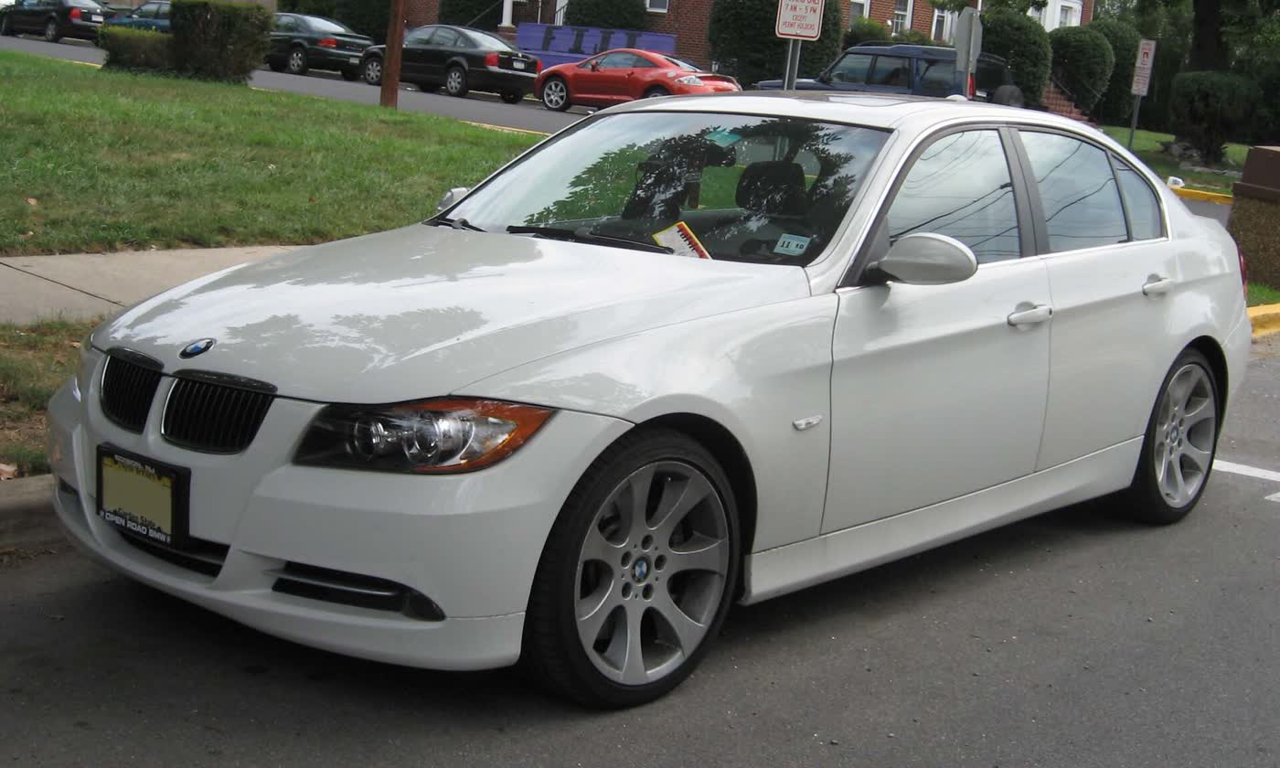 A white BMW 335i sedan of the first E90 style, front three-quarter view, parked at a curb on a residential street with grass behind it