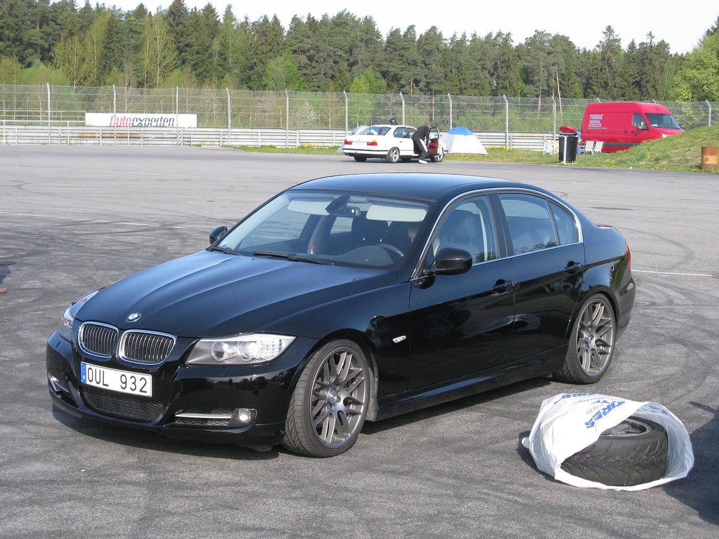 A black BMW 335i sedan with grey multi-spoke wheels parked on tire-marked tarmac, a wheel in a white bag beside it and a circuit fence behind