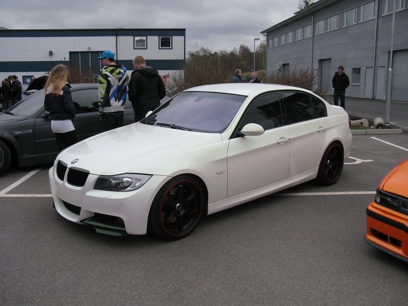 A white BMW 335i sedan on black aftermarket wheels with a deeper front bumper, parked at a car meet with people standing behind it