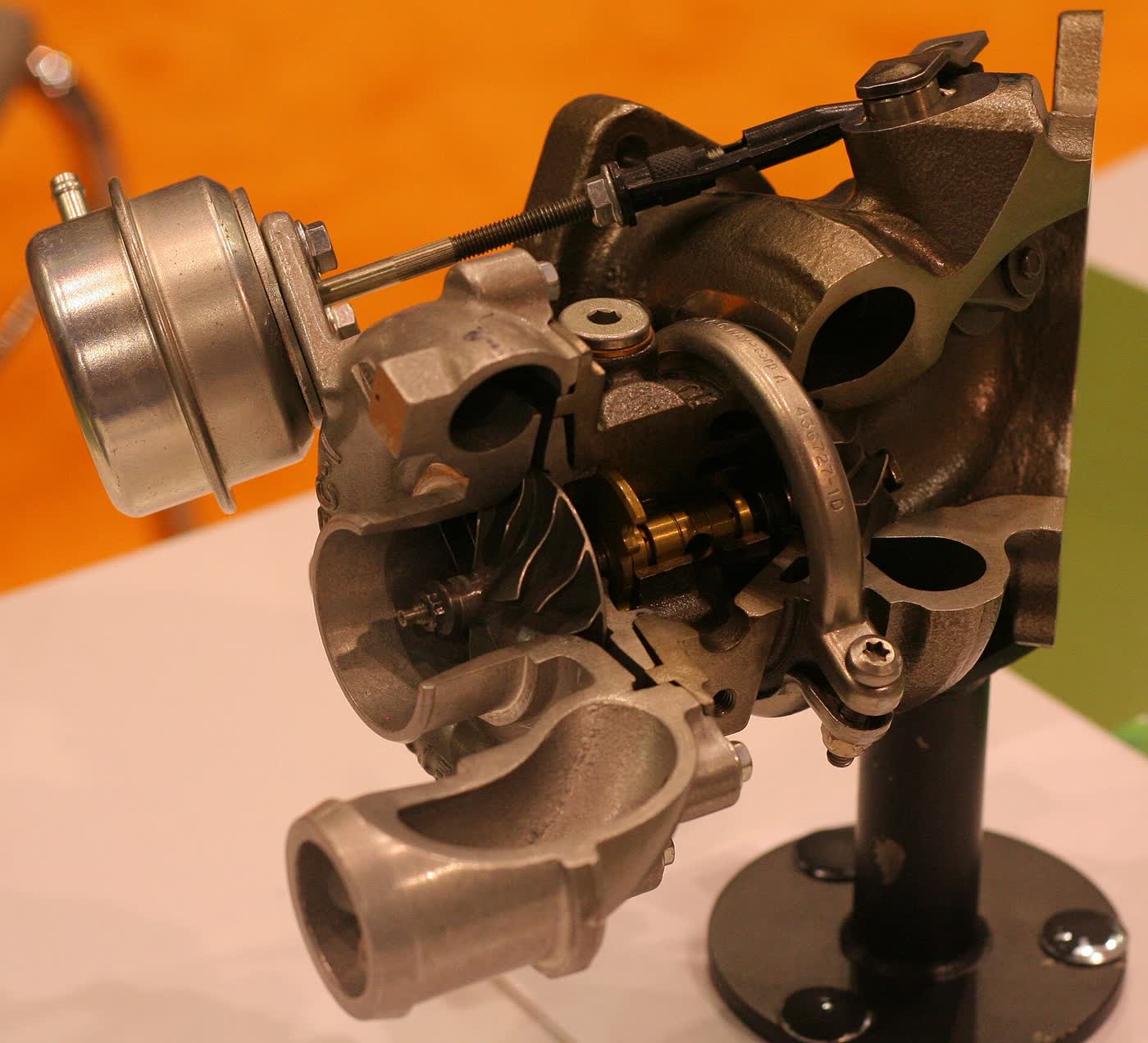 A cutaway turbocharger on a display stand, its housing sliced open to show the turbine wheel, with a round wastegate actuator and its rod mounted on top