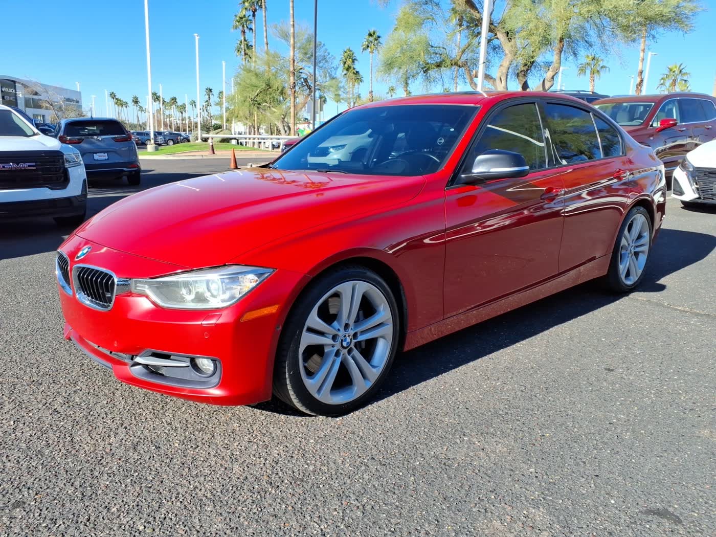 A red BMW 335i sedan with silver double-spoke alloy wheels, front three-quarter view, parked on a dealership lot with palm trees behind it