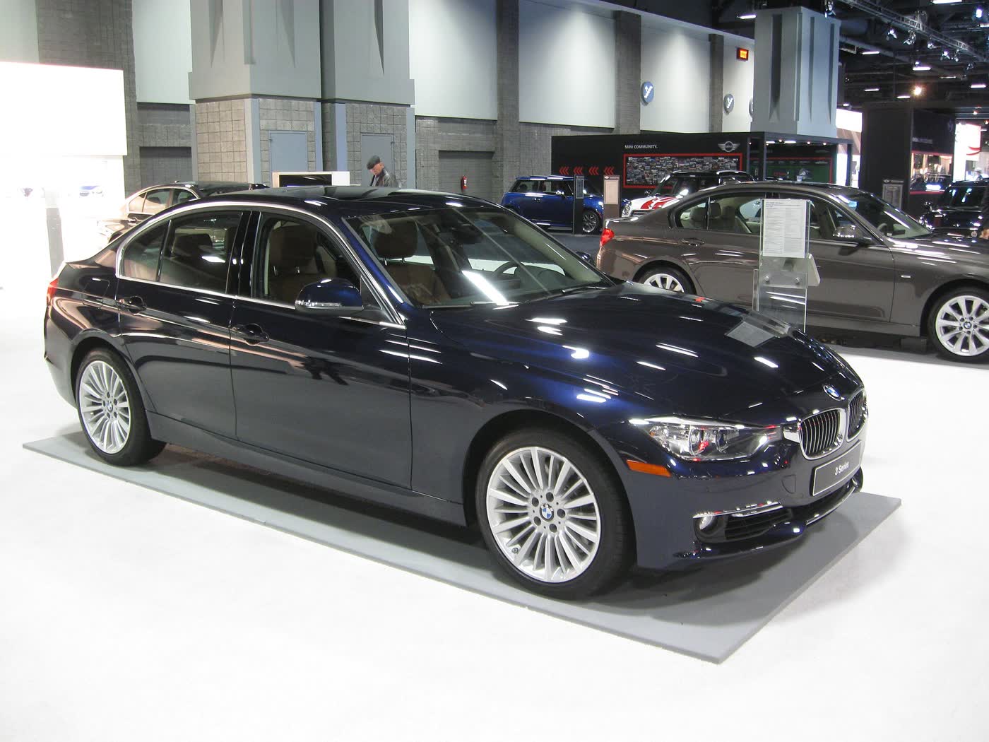 A dark blue BMW 335i sedan of the F30 generation with light multi-spoke alloy wheels, front three-quarter view, on a white display floor at an indoor car show