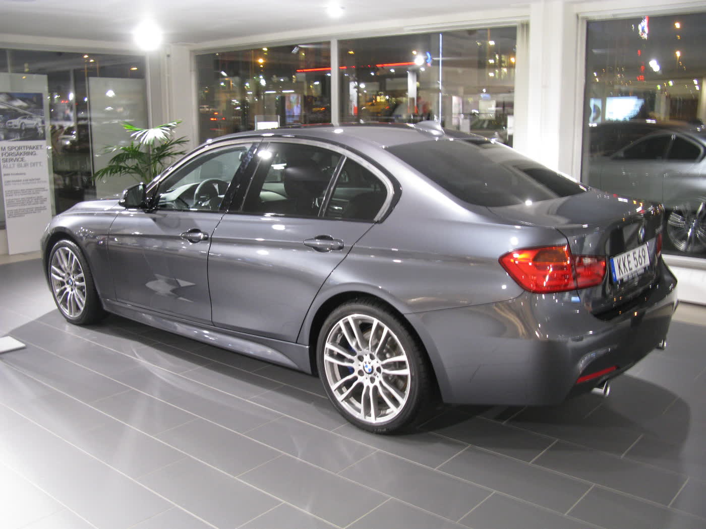 A grey BMW 335i M Sport sedan, rear three-quarter view, in a showroom with large silver alloy wheels and a Swedish number plate
