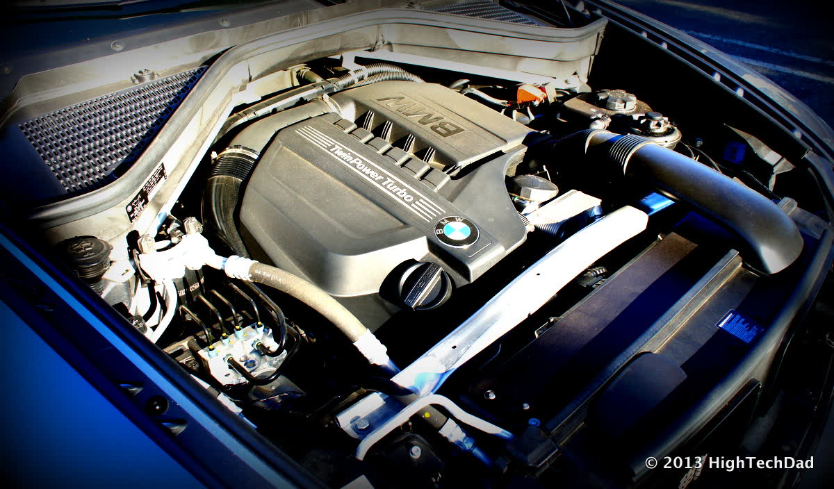 The engine bay of a BMW under the open hood, with a plastic engine cover reading TwinPower Turbo and the BMW roundel, a black intake duct on the right and a metal strut brace across the front