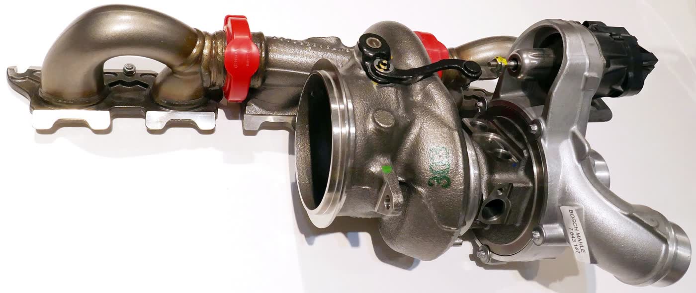 A BMW turbocharger removed from the engine and photographed on a white background, still bolted to its exhaust manifold section, with a black electric wastegate actuator at the right-hand end