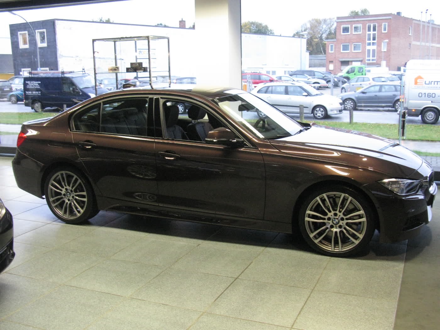 A dark brown BMW 335i sedan with multi-spoke alloy wheels, side view, parked in a showroom with a street visible through the window behind it