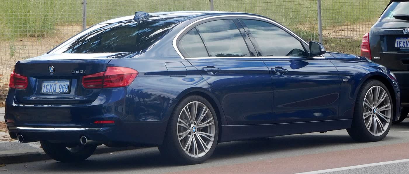 The rear of a dark blue BMW 340i sedan with a 340i badge on the boot lid, twin exhaust tips under the bumper and silver multi-spoke wheels, photographed kerbside in front of a wire fence