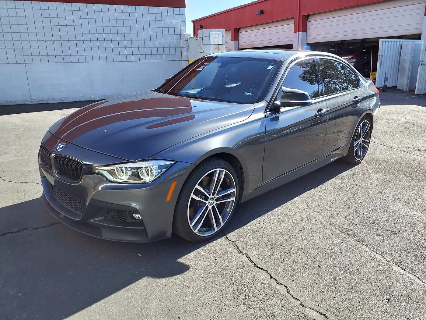 A dark grey BMW 340i sedan of the F30 generation with a front splitter and multi-spoke alloy wheels, front three-quarter view, parked on cracked asphalt in front of a red and white garage building