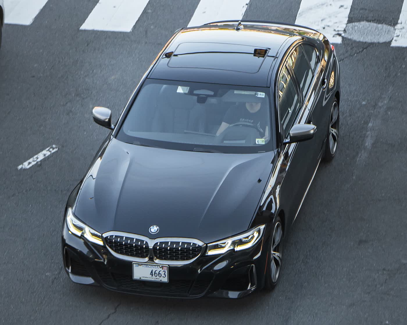 A black BMW M340i sedan seen from above at a crosswalk, its sunroof and the driver visible through the windshield