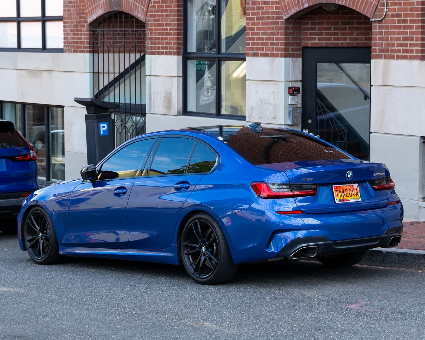 A blue BMW M340i sedan with black wheels, rear three-quarter view, parked at the curb in front of a brick building