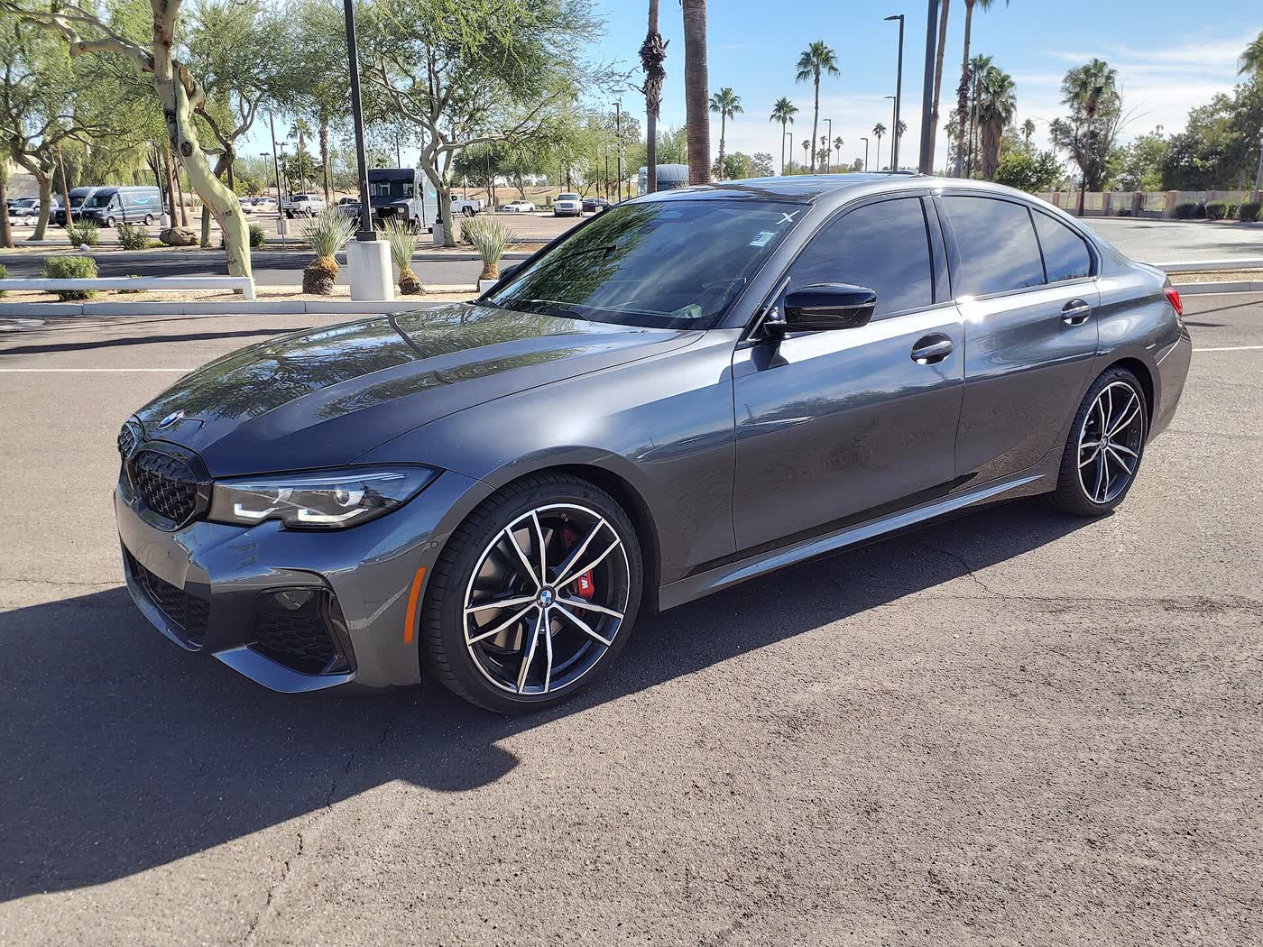A grey 2022 BMW M340i sedan with black-and-silver multi-spoke wheels and red brake calipers, front three-quarter view, parked on a dealer lot under palm trees