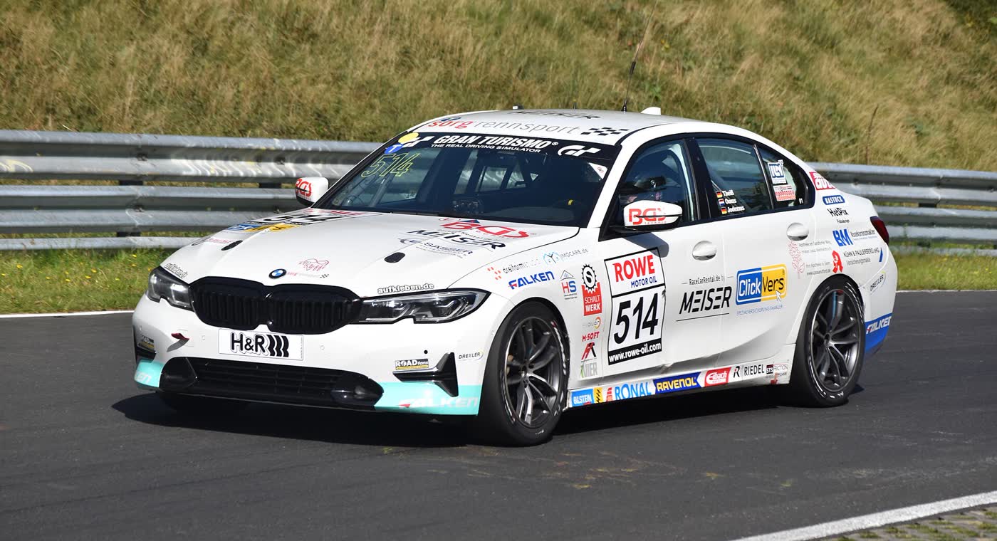 A white BMW 3 Series race car with sponsor decals and the number 514, cornering on a track with a guardrail and grass behind it