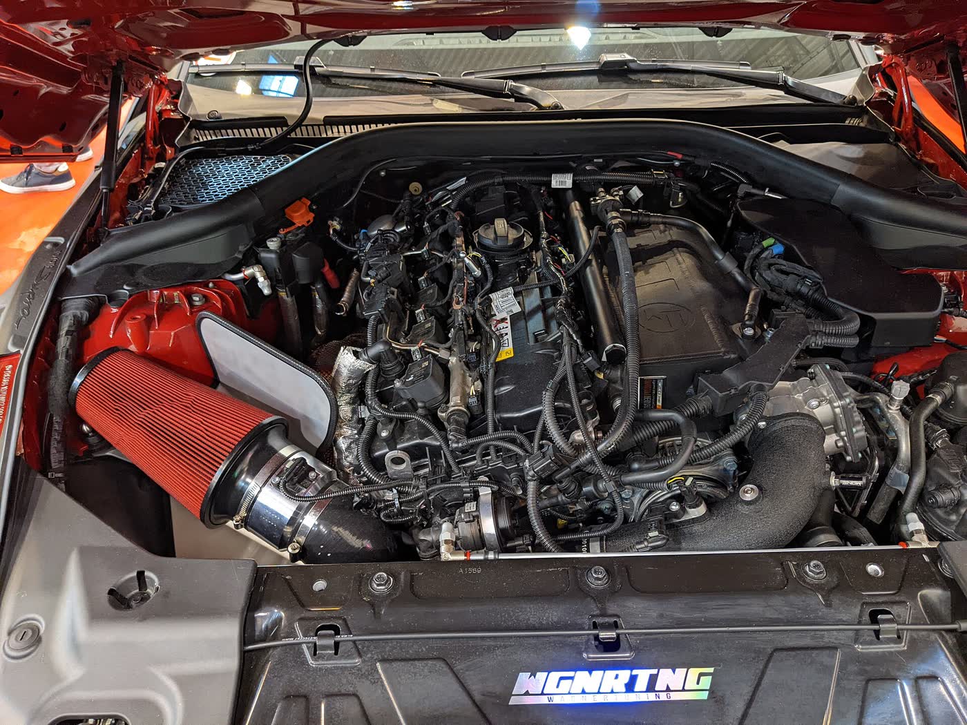 The engine bay of a red Toyota Supra with the cover off, a red cone air filter on an aftermarket intake at the left, and a tuner's illuminated logo on the front panel