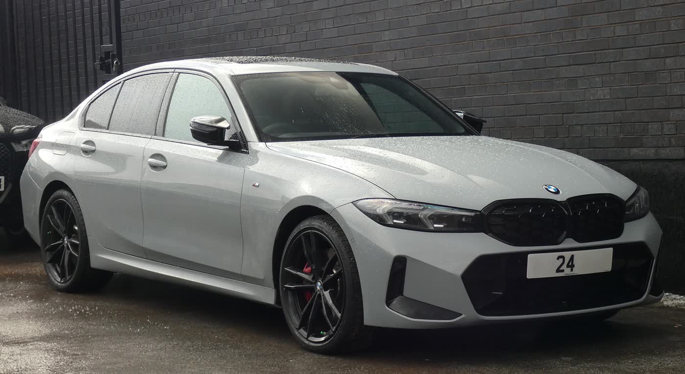 A light grey facelifted BMW M340i sedan with dark wheels and red brake calipers, parked against a dark brick wall on a wet day