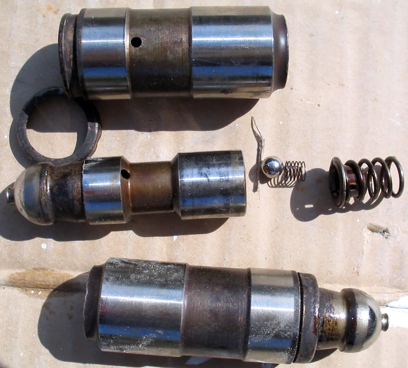 Three hydraulic valve lifters on a bench, one stripped to show its internal check ball and spring
