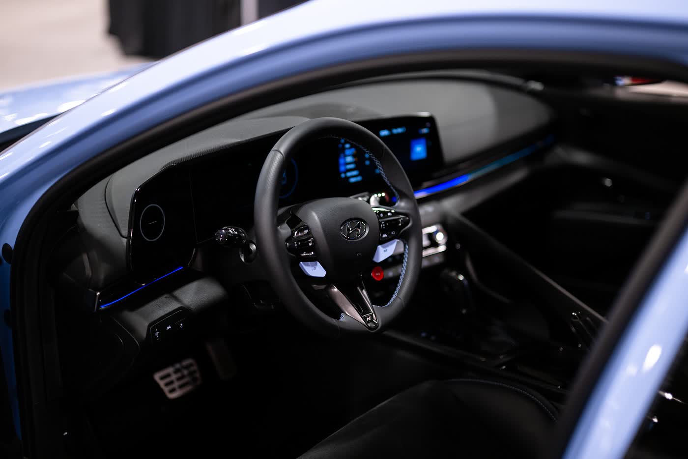The driver's side of a Hyundai Elantra N cabin seen through the open door: an N steering wheel with red buttons, and a digital gauge cluster and touchscreen in one curved housing