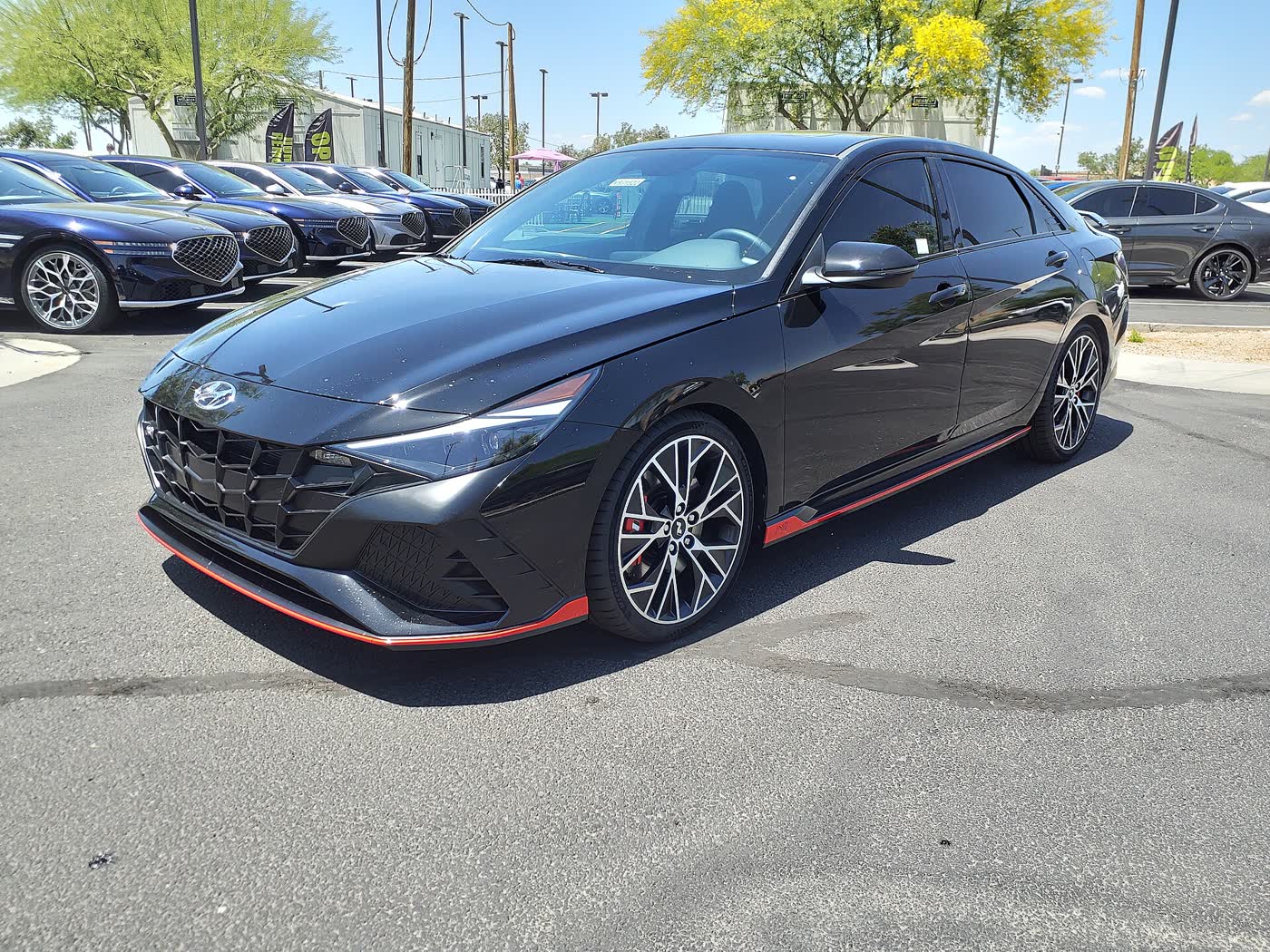A black 2023 Hyundai Elantra N sedan with red lower trim on a dealer lot, other cars parked in rows behind it
