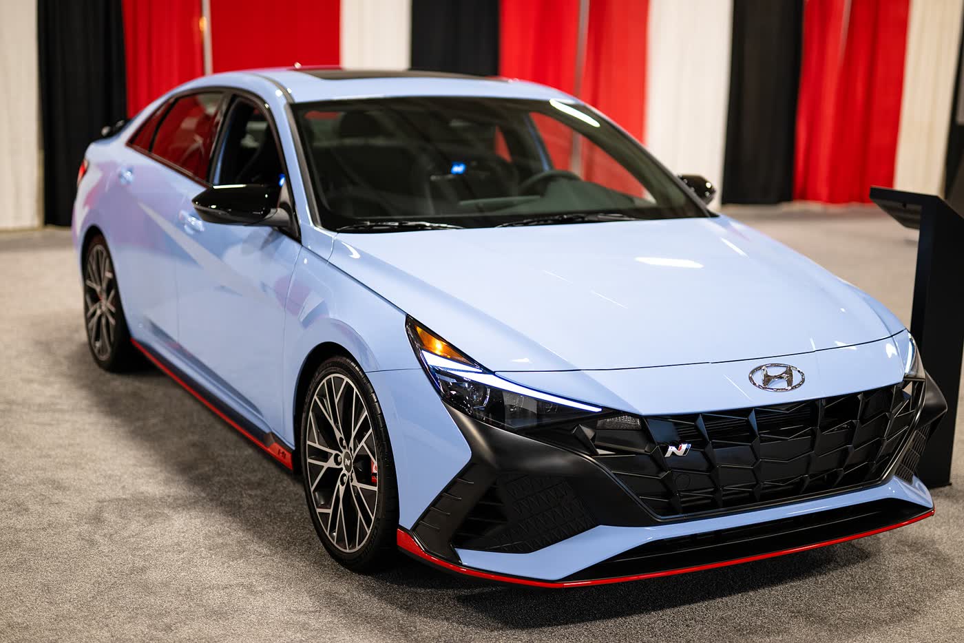 A light blue Hyundai Elantra N sedan with red lower trim and dark alloy wheels, front three-quarter view, on an auto show stand