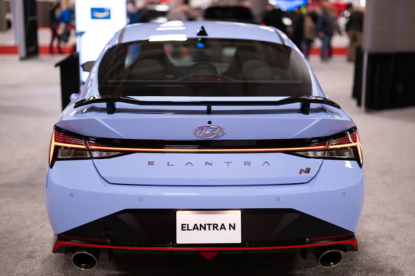 The rear of a light blue Hyundai Elantra N on an auto show stand: a wing across the trunk lid, a black diffuser and two large exhaust tips