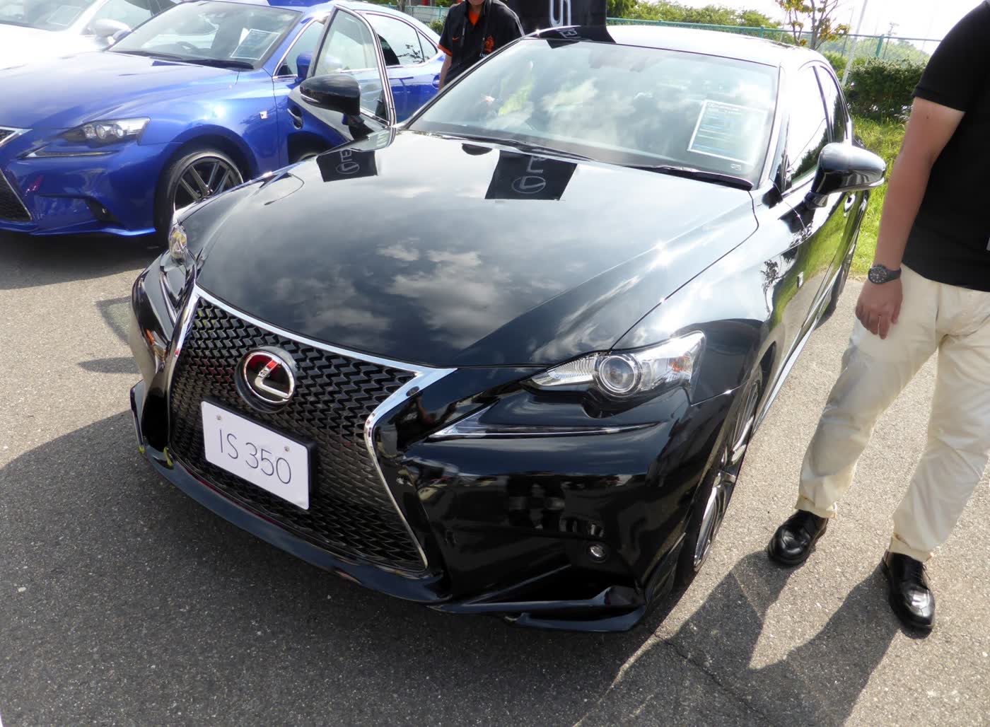 A black Lexus IS 350 F Sport sedan with a mesh grille and an IS 350 plate, seen from the front left at a car meet, with a blue IS beside it and a man standing at the right edge