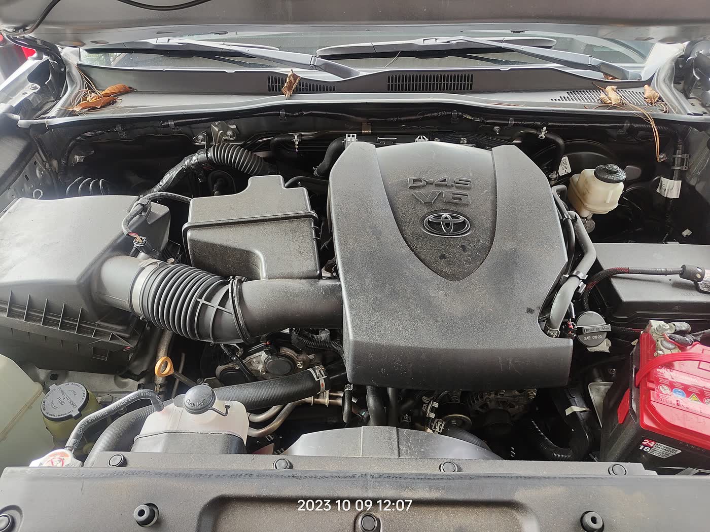 The engine bay of a Toyota Tacoma with its black plastic engine cover reading D-4S V6, an air intake duct on the left and a red battery on the right