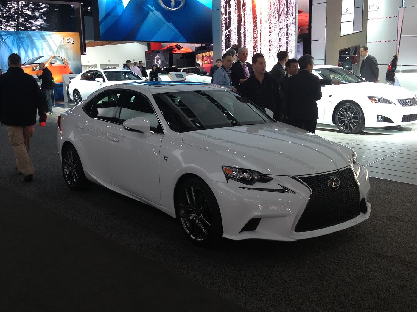 A white third-generation Lexus IS 350 F Sport sedan on a show-floor stand, front three-quarter view, with its black mesh grille and black multi-spoke wheels, visitors standing behind it