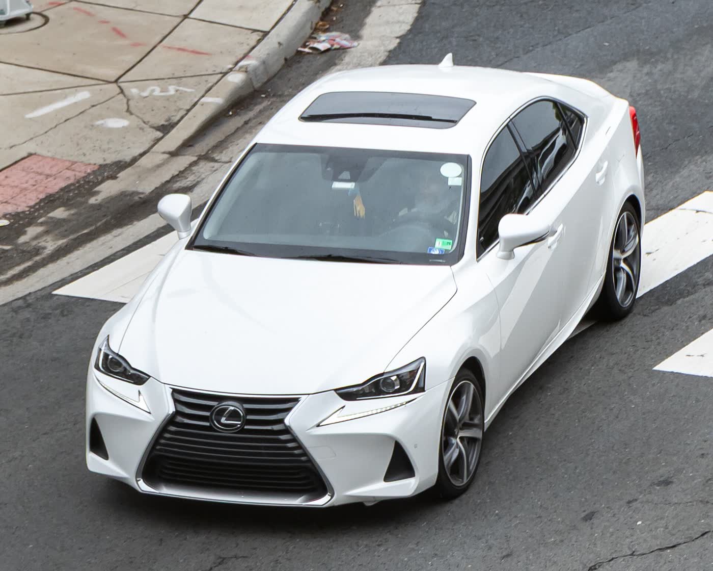 A white Lexus IS sedan with a sunroof, seen from above and ahead as it crosses a painted crosswalk, with the refreshed headlamp and grille styling
