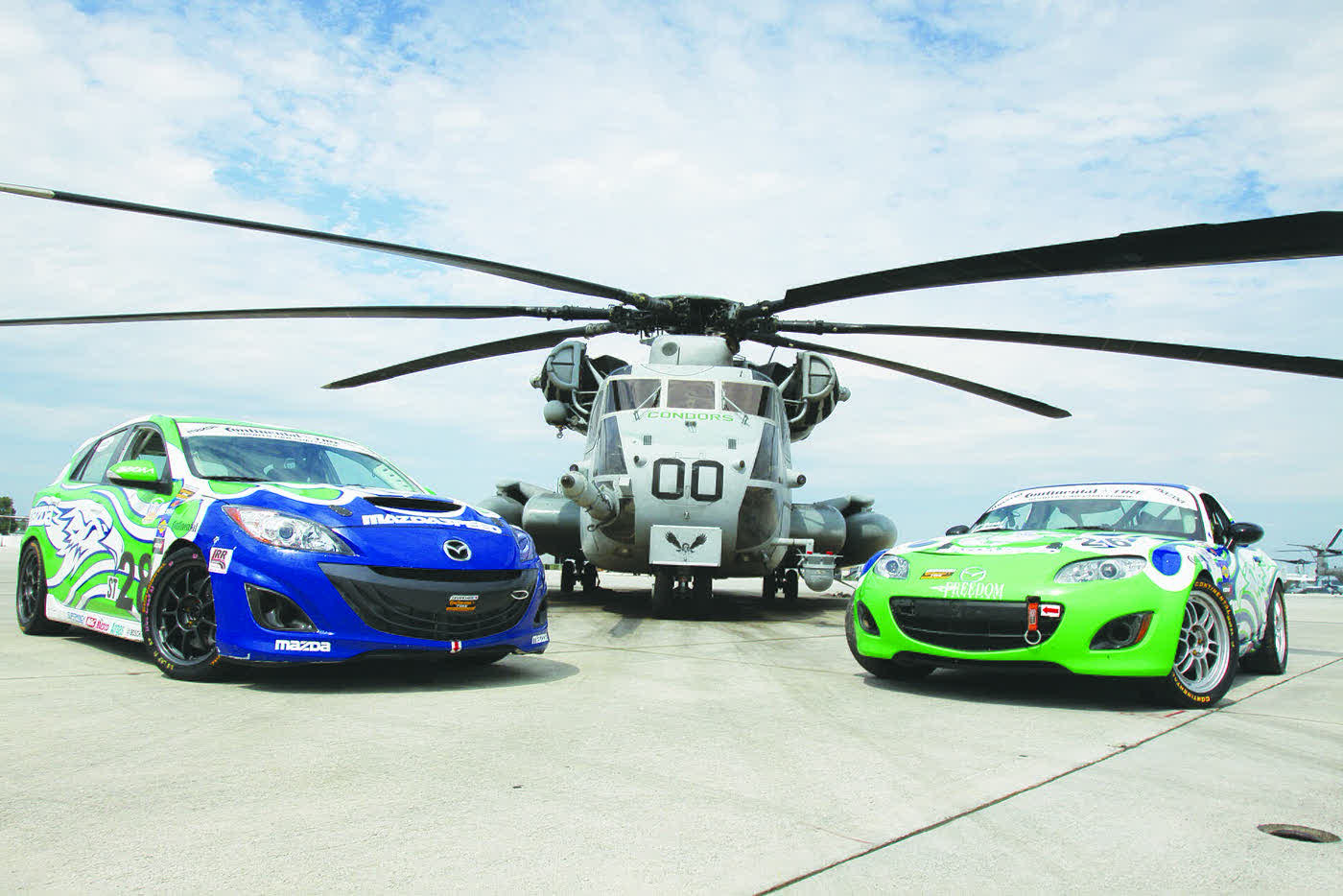 A Mazdaspeed3 in Grand-Am race livery parked beside an MX-5 race car in front of a military helicopter