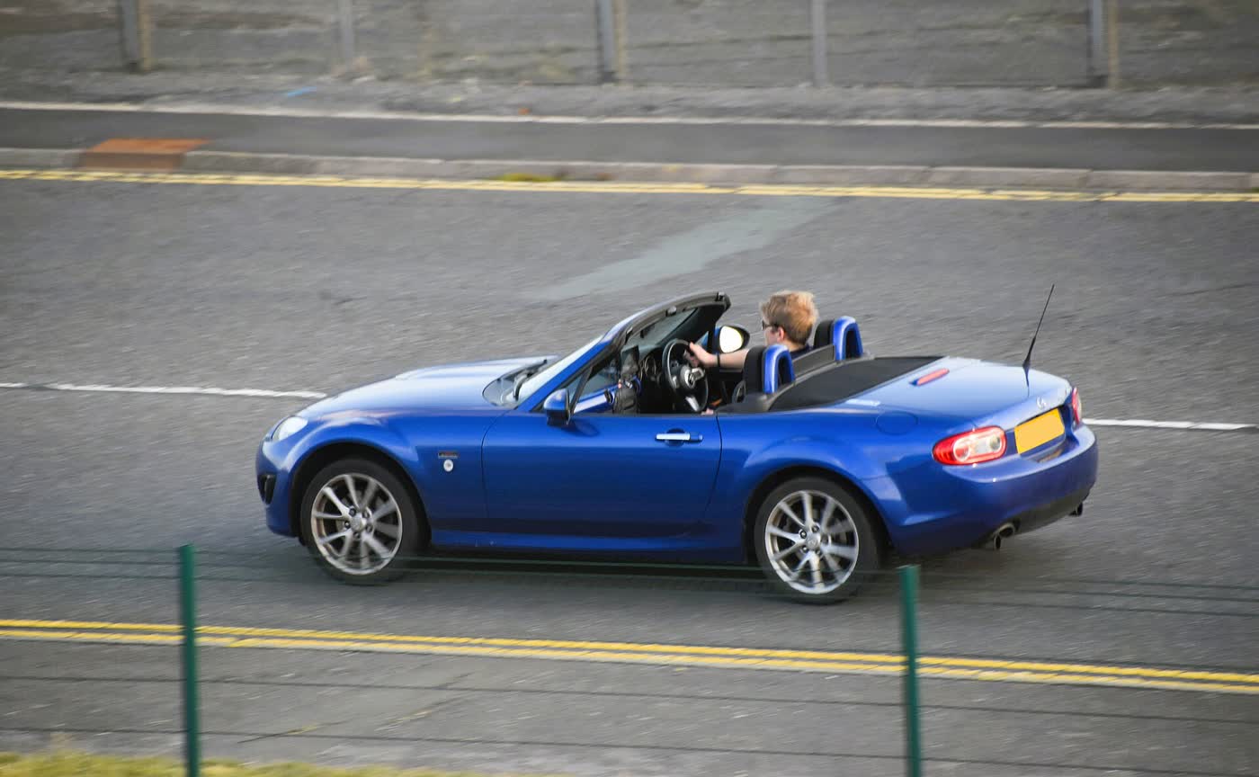 A blue Mazda MX-5 NC being driven on a road with the roof down