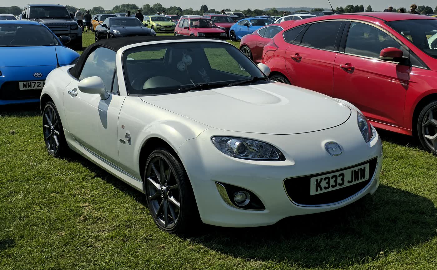 A white 2012 Mazda MX-5 NC with its black soft-top raised, parked on grass at a car meet