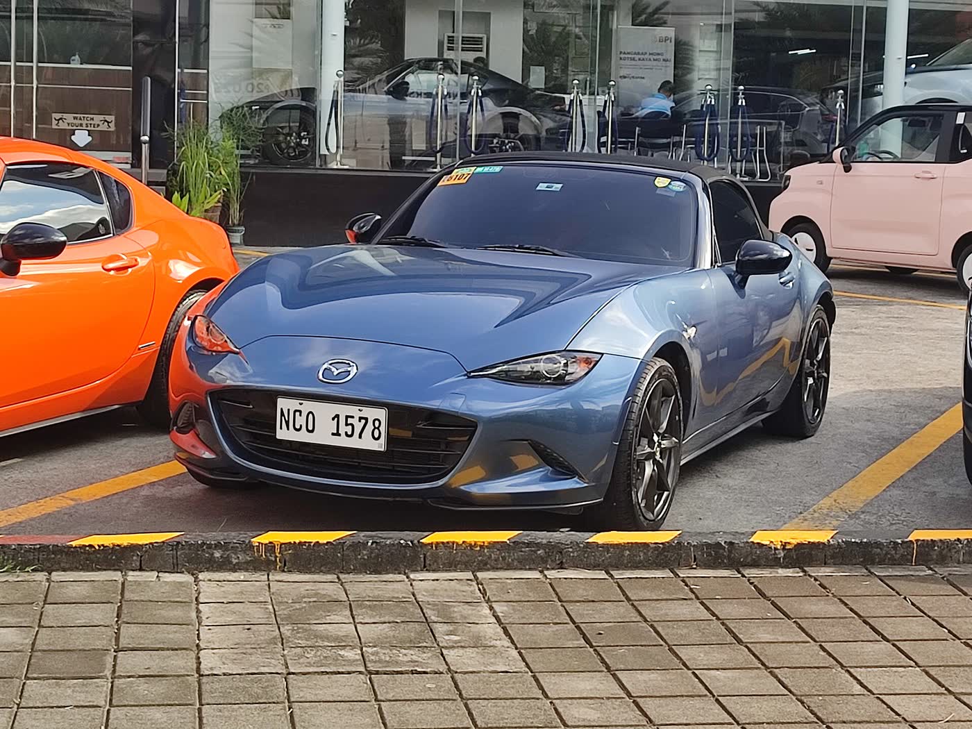 A blue Mazda MX-5 ND parked outside a Mazda dealership with window stickers still in the glass
