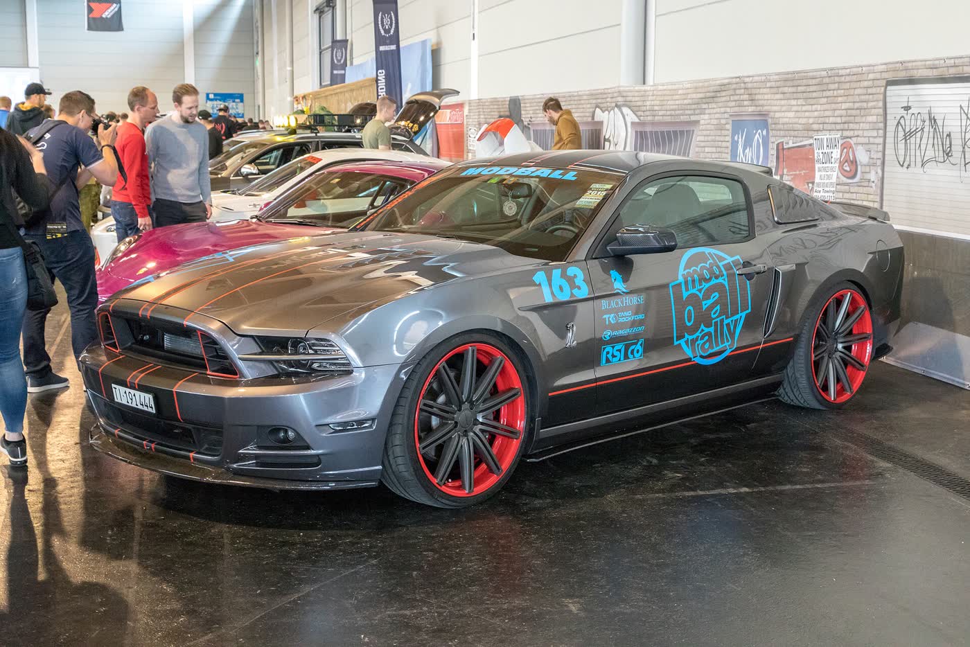 A grey modified Ford Mustang S197 on aftermarket wheels with rally livery, displayed at a tuning show