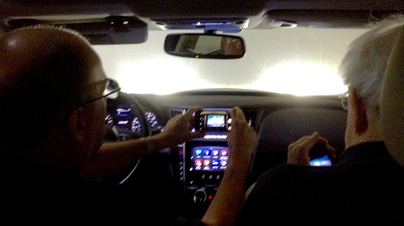 Two passengers in the front of an Infiniti Q50 at dusk, one photographing the upper and lower dashboard touchscreens