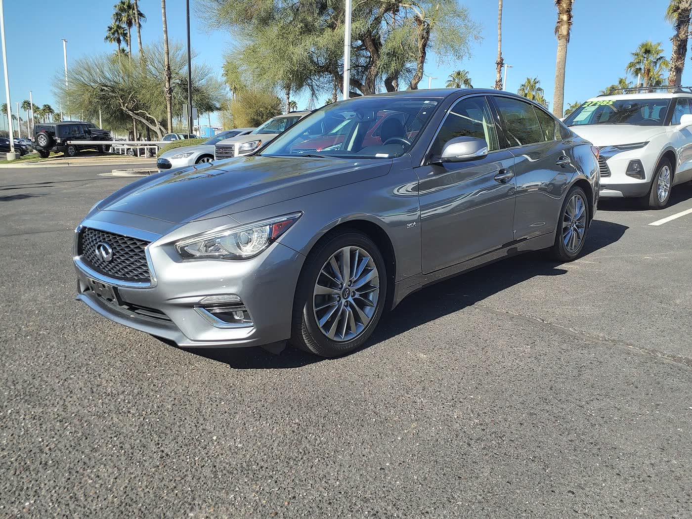 A grey 2018 Infiniti Q50 sedan with a 3.0t badge on its front fender, front three-quarter view, parked on a dealer lot under a clear sky