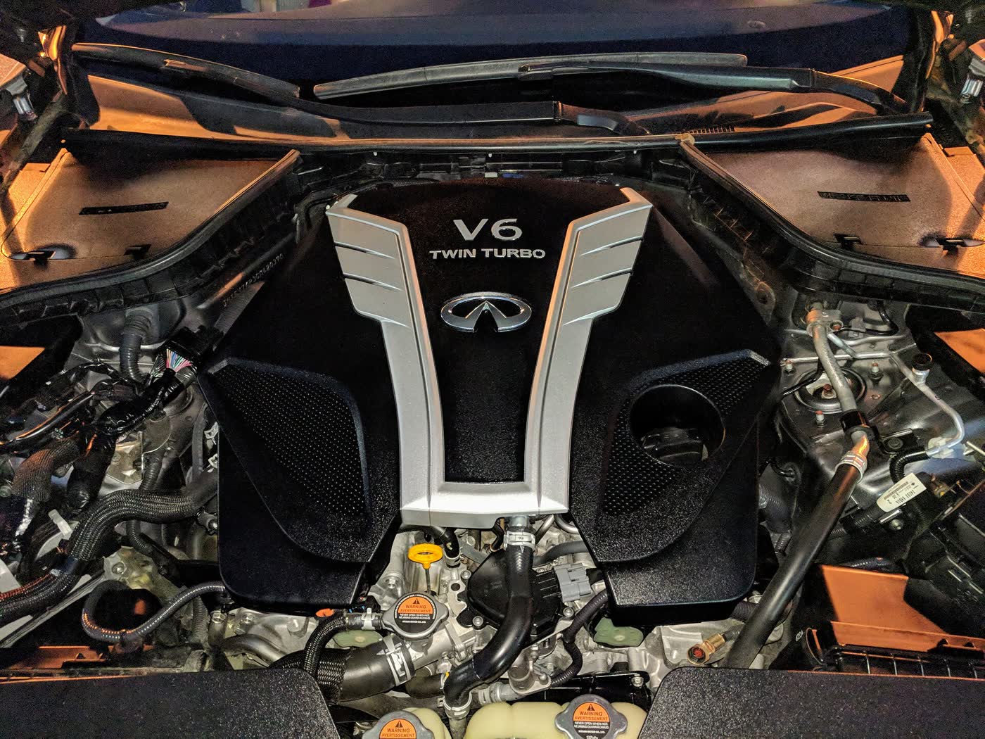 An engine bay seen from above, with a silver and black V6 TWIN TURBO engine cover carrying the Infiniti badge